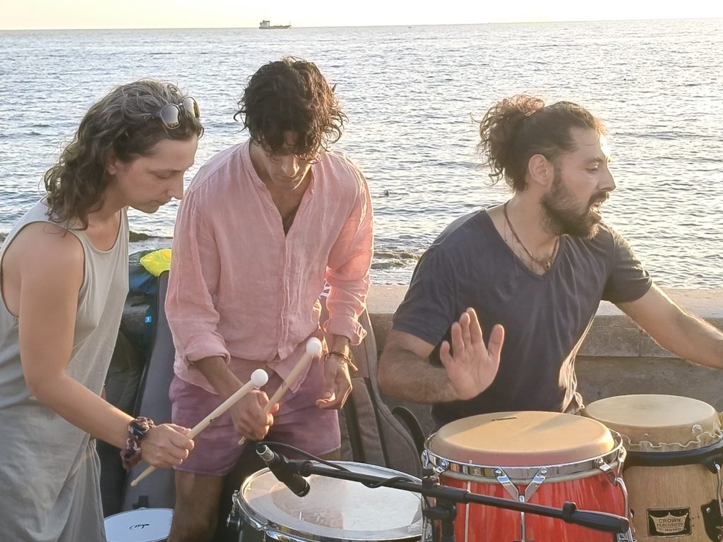 Tre percussionisti del Colectivo con rullante e congas davanti al mare, nella luce calda del tardo pomeriggio