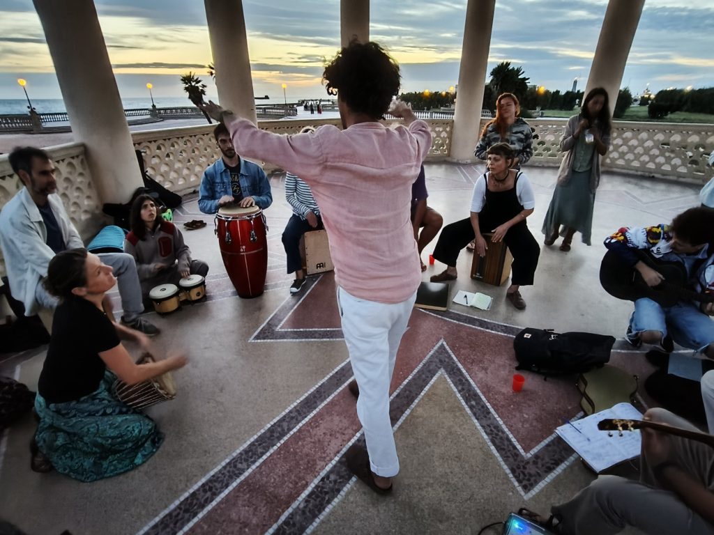 Il Colectivo in cerchio sotto il gazebo della Terrazza Mascagni a Livorno al tramonto, con il direttore di spalle che dà l'attacco e il mare sullo sfondo